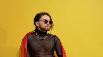 Gay man dressed in mesh t-shirt and sunglasses sits on yellow background with a multicolored rainbow flag. Concept diversity, transsexual, and freedom video