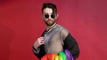 gay uomo vestito nel maglia maglietta e occhiali da sole su rosso sfondo con un' multicolore arcobaleno bandiera. concetto diversità, transessuale, e la libertà video