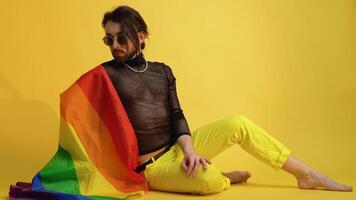 Gay man dressed in mesh t-shirt and sunglasses sits on yellow background with a multicolored rainbow flag. Concept diversity, transsexual, and freedom video