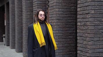 een jong Kaukasisch elegant Mens wandelingen Aan de straat in een jas Holding een geel sjaal in zijn hand- video
