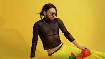 Gay man dressed in mesh t-shirt and sunglasses sits on yellow background with a multicolored rainbow flag. Concept diversity, transsexual, and freedom video