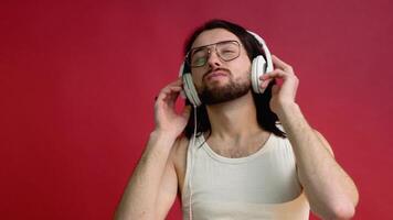 jovem sorridente feliz alegre metrosexual homem dentro óculos e branco camiseta ouvindo para música em vermelho cor fundo video