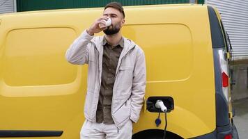 A man drinks coffee while leaning on his cargo electric car, standing on the charging station for charging a car. Electric car charging concept video