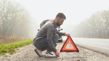 homem colocando triângulo Cuidado placa em nebuloso estrada perto quebrado carro video