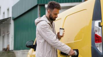 Español hombre enchufar en cargando cable a eléctrico vehículo y cargos pilas un hombre bebidas café mientras propensión en su carga eléctrico coche video