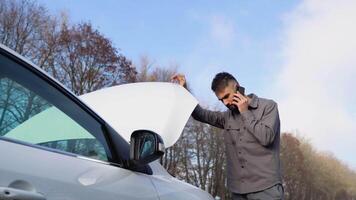 Sad upset guy driver is standing near broken car, calling emergency on cell mobile phone talking on smartphone. Young desperate man got into accident video