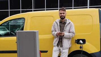 sorridente latim homem dentro casual vestem com telefone enquanto inclinado em dele carga elétrico carro, em pé em a cobrando estação para cobrando uma carro. elétrico carro cobrando conceito video