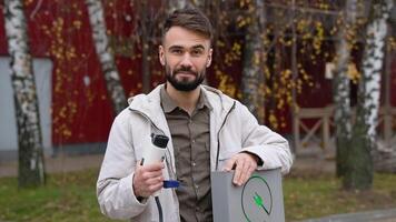 A man with a charger at a charging station looks into the camera. Electric car charging concept video