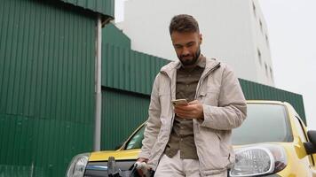 Stylish handsome male standing near modern electric car and texting on smartphone outddors. Young man in good mood browsing on cellphone. Urban concept video