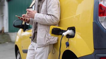 joven hombre chateando en móvil teléfono mientras eléctrico coche es cargando en al aire libre estación, cerca arriba video