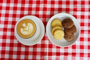Choco chip cookies and latte coffee on a white plate with a checkered tablecloth seen above photo