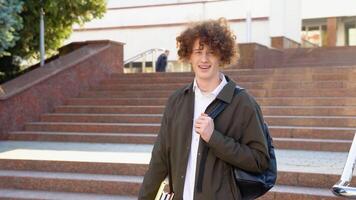 Outdoor portrait of cheerful red -haired curly student with backpack and books standing near college building, looking at camera and smiling video