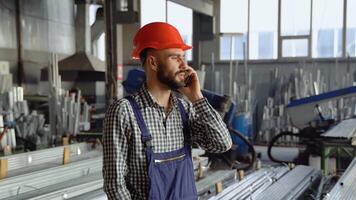 A young worker in a helmet at a large metalworking plant speaks on the phone. Professional worker in the finished product warehouse makes an order video