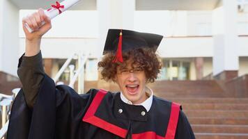 rouge -aux cheveux frisé gars avec un appareil dentaire des stands dans une maîtrise manteau et triomphalement soulève le sien diplôme plus de le sien tête video