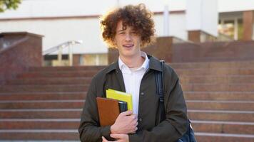 Outdoor portrait of cheerful red -haired curly student with backpack and books standing near college building, looking at camera and smiling video