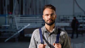 Portrait of bearded factory worker in protective glasses looking at camera while standing in workshop video