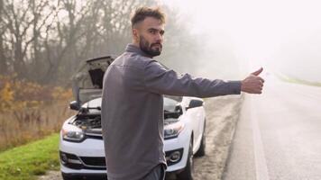 Upset young man hitchhiking standing near a broken car at foggy roadside video