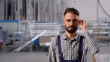 Portrait of bearded factory worker in protective glasses looking at camera while standing in workshop video