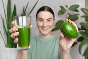 mujer sonriente y muestra verde hierba de trigo bebida y aguacate. sano estilo de vida concepto, verde sano vegetales y frutas foto