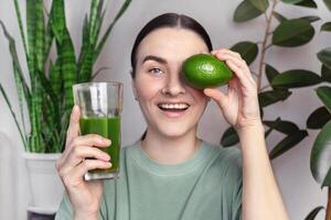 mujer sonriente y muestra verde hierba de trigo bebida y aguacate. sano estilo de vida concepto, verde sano vegetales y frutas foto