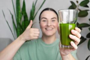 mujer sonriente muestra un pulgares arriba. ella Bebiendo verde hierba de trigo energía agitar. foto