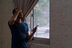 joven asiático hombre utilizando teléfono inteligente estresado con mano en cabeza junto a de ventana foto