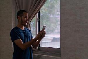 emocionado joven asiático hombre en pie junto a de ventana con inteligente teléfono foto
