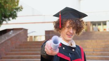 une Jeune rouge -aux cheveux frisé gars avec un appareil dentaire des stands Extérieur dans une maîtrise manteau et étend une diplôme vers l'avant à caméra video