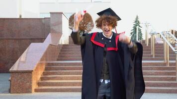 A young curly funny graduate in the master's mantle rejoices in completing his studies and bouncing with a diploma in his hands video