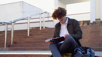 Jeune rouge -aux cheveux frisé étudiant est assis sur le escaliers lit livres dans une Université Campus video