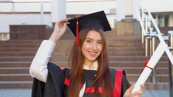 glimlachen rood -harig gekruld meisje met een beugel staat in een meester mantel en Holding Aan naar een hoed en een diploma video