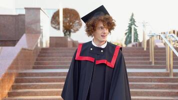 A young curly funny graduate in the master's mantle is having a master's clothing. Curly graduate with braces smiles near the college video