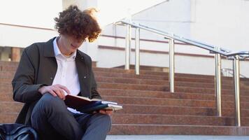 jong rood -harig gekruld leerling zit Aan de trap leest boeken in een Universiteit campus video