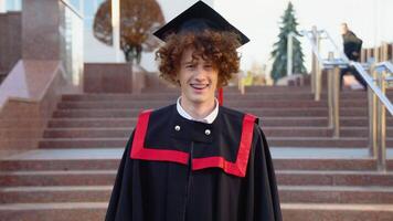 A young funny graduate in the master's mantle smiles at the camera. Curly graduate with braces smiles near the college video