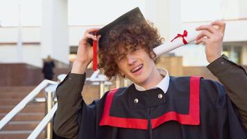 Happy red -haired curly guy with braces stands in a master's mantle and holding on to a hat and a diploma video