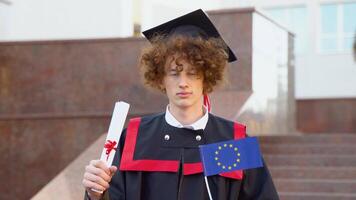 A curly funny smiling graduate in the master's mantle holds a diploma of completion of his studies and the flag of the European Union and yawns video