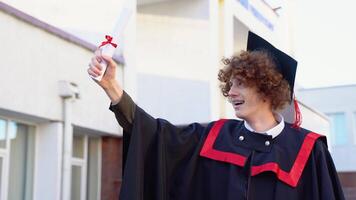 baixo ângulo retrato do feliz triunfante masculino graduado em pé perto universidade segurando acima diploma. a partir de abaixo do jovem bonito homem orgulhoso do acadêmico conquistas a comemorar Faculdade graduação video