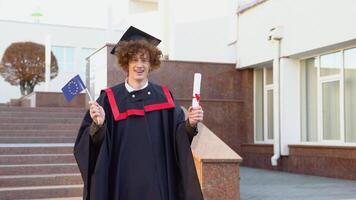 A young curly funny graduate in the master's mantle holds a diploma of completing his studies and the EU flag video
