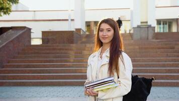 Cheerful student girl hugging books posing with backpack near college building outdoor, smiling to camera. Modern education, college tuition and grants, studentship and study abroad concept video