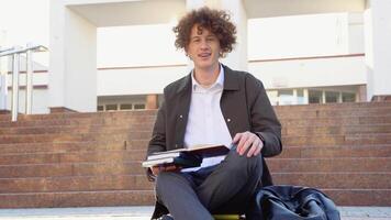 contento hermoso exitoso estudiante con tirantes, se sienta en el pasos y sonriente, mira dentro el marco al aire libre en el calle cerca el Universidad video