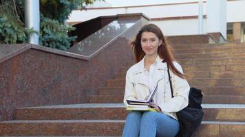 contento alunno ragazza in posa con zaino Tenere libri sorridente guardare a telecamera seduta su il le scale vicino Università edificio all'aperto. moderno formazione scolastica e borsa di studio stile di vita video