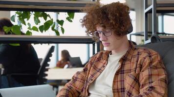 Young redhead blogger or freelancer with headphones holding notebook browsing chatting while sitting on comfy soft armchair wears yellow shirt in co working space video