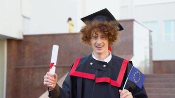 A curly funny graduate in the master's mantle holds a diploma of completion of his studies and the flag of the European Union and yawns video