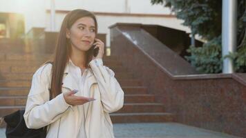 Cheerful student girl hugging books posing with backpack near college building outdoor, talking on the phone. Studentship and study abroad concept video