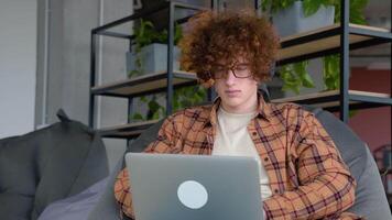 Young redhead man with headphones holding notebook browsing chatting while sitting on comfy soft armchair wears yellow shirt in co working space video