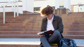 giovane rosso -dai capelli Riccio alunno si siede su il le scale legge libri nel un' Università città universitaria video