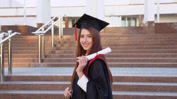 ein Universität weiblich Absolvent steht im ein Meister Mantel und elegant hält ein Diplom auf ihr Schulter video