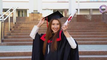 A university female graduate stands in Master's mantle holds a master's hat and triumphantly waved a diploma over her head video