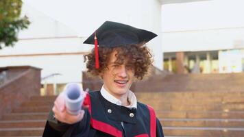 ein jung rot -behaart lockig Kerl mit Hosenträger steht im ein Meister Mantel halten ein Diplom gedehnt nach vorne zu Kamera video