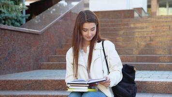 joven mujer estudiante es leyendo libro cerca Universidad edificio. niña es estudiando, preparando a examen a Universidad instalaciones. educación concepto. alto colegio estilo de vida video